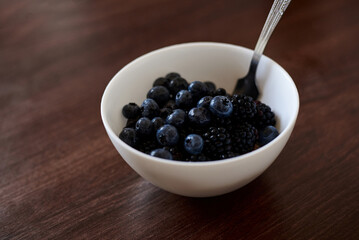 A white bowl filled with dark blue blackberries isolated on brown wooden table. Healthy nutrition ration. Fruit and vegetable diet. Raw food. Eat healthy, keep fit.