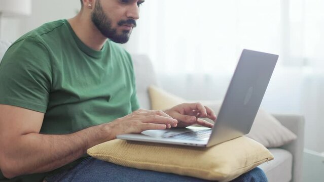 Focused indian freelancer sitting on comfy couch and typing on wireless laptop. Bearded guy in casual wear using yellow soft pillow on knees as support for portable computer.