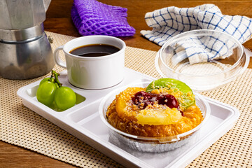 High angle view of delicious homemade mixed fruit danish, a cup of black coffee and grapes in white plastic tray on table mat on wooden table.