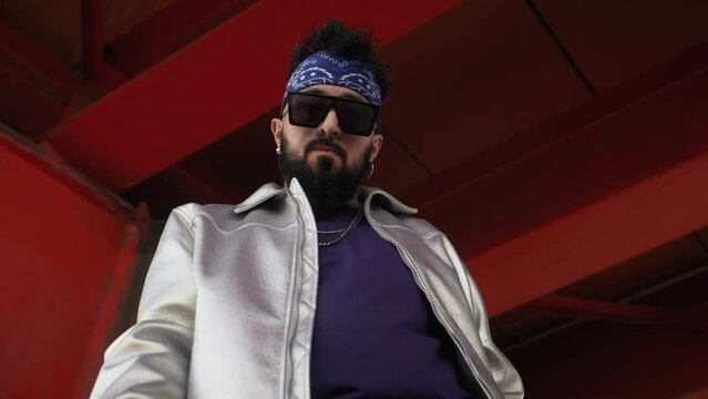 Low-angle shot of rapper in shiny jacket and sunglasses showcasing rap hand gestures on city street