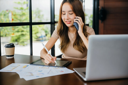 Asian businesswoman sitting at cafe calling mobile phone talking with customer asking about something, smiling female working laptop computer and writing take note on digital tablet at home office - Powered by Adobe