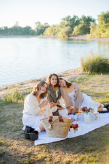 Young beautiful women of 25 years old on an autumn picnic near the lake. Glass of white wine, pastries. Happy models chatting merrily. Sunset.