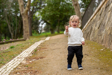 Cute blonde kid with a stick is playing around in a park. Happy adorable caucasian baby boy attacking with a stick. A boy in a white sweater pointing with a stick at the camera.