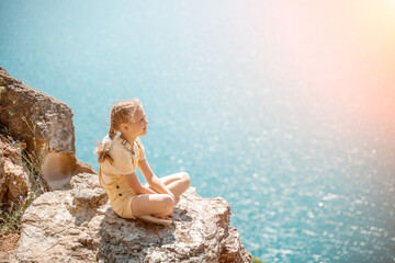 Happy girl perched atop a high rock above the sea, wearing a yellow jumpsuit and braided hair,...