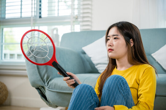 Asian female sitting floor using mosquito device swatter or electric net racket, Young woman killing mosquitoes hand holding fly swatter like weapon in living room at home, technology insect killer