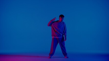 A young man stands against a blue background and looks at the camera. A pink light shines on him from the side and illuminates one side. He is a dancer performing a dance and showing the moves