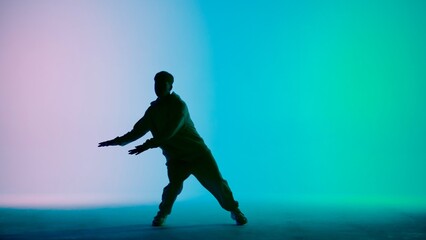 In the frame on a tricolor background, gradient in silhouette stands a young man. Demonstrates dance moves, stretching his arms forward. He is plastic, rhythmic. He is dressed in street style clothes