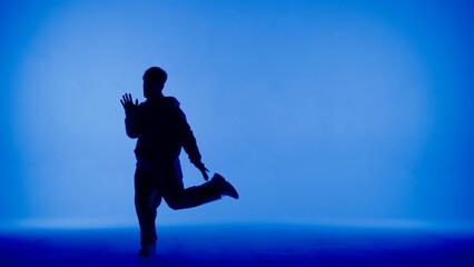 Fototapeta na wymiar In the frame on a blue and white background, a young man stands in silhouette. Demonstrates dance movements, raising one leg. He is plastic, rhythmic. He is dressed in street style clothes.