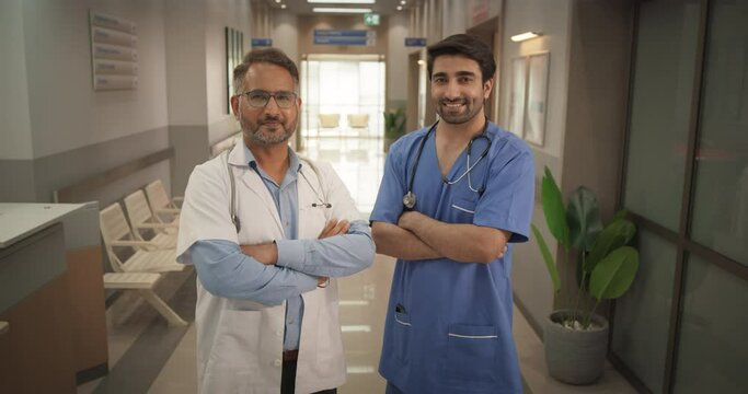 Two Friendly Indian Doctors: Team Portrait of Male Successful Medical Healthcare Professionals Standing in a Modern Hospital Office, Posing, Looking at Camera and Smiling. Doctor and Nurse on Duty