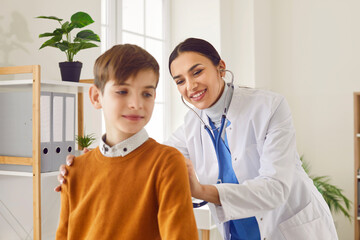 Happy, smiling woman doctor pediatrician in a white medical coat uses a stethoscope to check heart beat or respiration of a little school child boy from his back. Health checkup concept