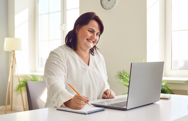Smiling overweight young woman using laptop in office. Happy plus size woman reviewing financial documents and making notes in copybook. Financial manager, bookkeeper, accountant - obrazy, fototapety, plakaty