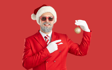 Man in Christmas costume showing Christmas tree ball. Happy bearded man in red suit, Xmas hat and...
