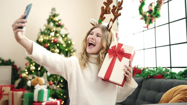 Young blonde woman making selfie by smartphone holding christmas gift at home