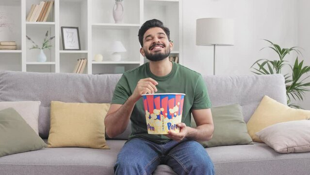 Handsome bearded guy of indian nationality sitting on cozy sofa. Young man enjoying tasty popcorn and watching TV with real interest. Concept of people, leisure and entertainment.