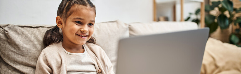 happy elementary age girl sitting on sofa and using laptop in modern living room, e-learning banner