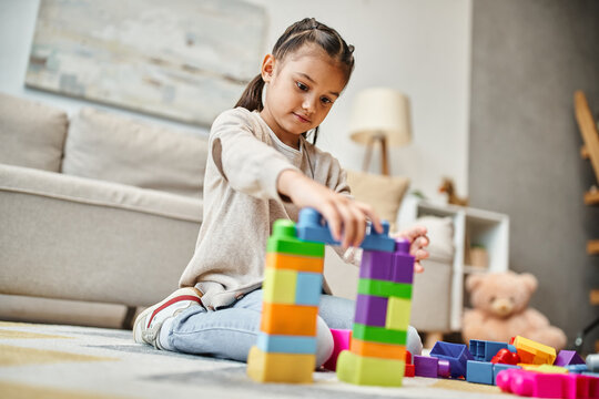 cute girl playing with colorful toy blocks on carpet in modern living room, building tower game