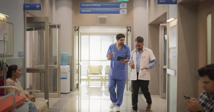 Slow Motion Portrait of Two Indian Male Doctors Walking in Hospital Corridor and Talking while Using a Digital Tablet. Two Medical Specialists Discussing the Treatment of a Patient
