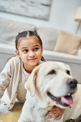 happy cute girl sitting on carpet and stroking dog in modern living room, kid and labrador