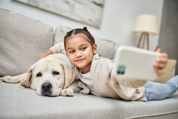 cute elementary age girl taking selfie with labrador dog in modern apartment, domestic animal