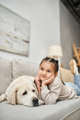 cheerful girl smiling and lying on sofa with cute labrador in modern living room, pet and child