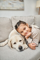 portrait of happy kid smiling near cute labrador in modern living room, furry friend and child
