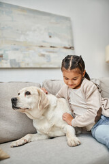 joyful girl sitting on sofa and cuddling cute labrador in modern living room, animal companion