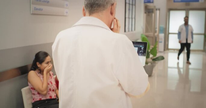 Backview Tracking Shot of Indian Doctor Walking in Hospital Corridor, Using a Digital Tablet. Slow Motion of Senior Male Surgeon Checking Brain MRI Images Before Medical Tumor Removal Intervention