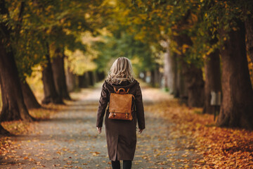 Stylish woman with brown trench coat and backpack walks in alley at autumn park. Beauty and fashion lifestyle