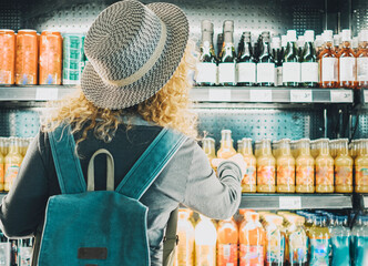 Back view of woman tourist with backpack in front of a window store full of drinks bottles choosing...