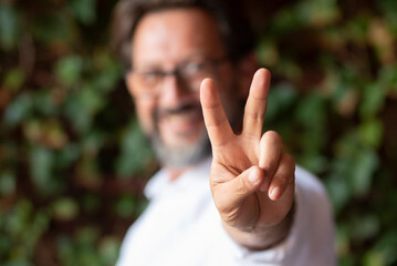 Focus on finger doing victory and peace gesture with cheerful and happy adult man in background. Green nature outdoor leaves defocused. Peaceful people and hope concept
