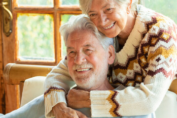 Happy and enjoyed senior caucasian couple smile and hug each other sitting on a chair in cozy home...