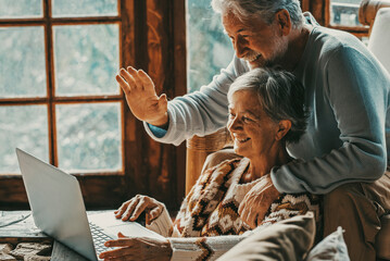 Happy senior couple man and woman doing video call with laptop at home sitting on the floor and chair together having un. Hallo gesture and modern mature people with connection technology leisure - obrazy, fototapety, plakaty