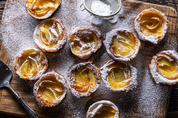 Mini tarts made of puff pastry and sliced apples sprinkled with powdered sugar on wooden cutting board - obrazy, fototapety, plakaty
