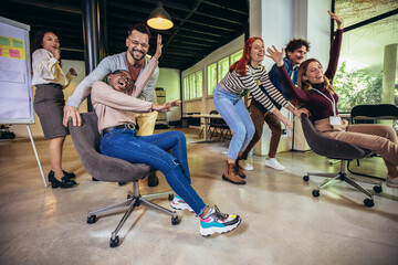 Young cheerful businesspeople in smart casual wear having fun while racing on office chairs and...