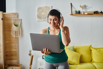 Fototapeta na wymiar cheerful short haired woman with headphones looking at laptop camera during online fitness lesson