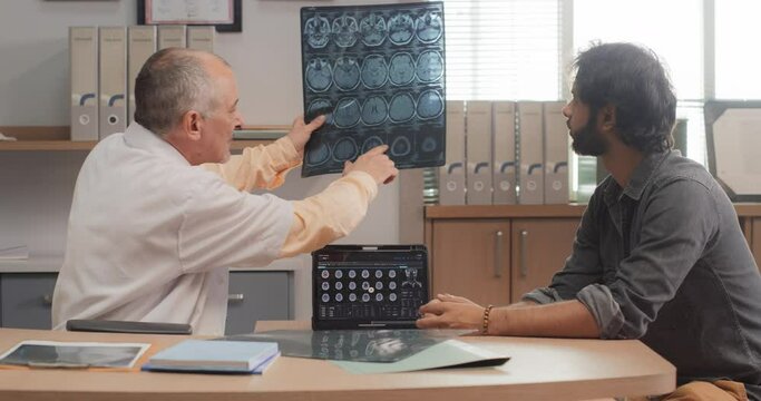 Hospital Ward: Middle-Aged Indian Male Doctor Talks to his Patient During Medical Appointment, Shows and Explains Brain CT MRI Scan Results. Physician Talks to a Man Before Surgery