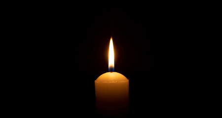 Single burning candle flame or light glowing on a big white candle on black or dark background on table in church for Christmas, funeral or memorial service with copy space. - obrazy, fototapety, plakaty