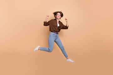 Fototapeta na wymiar Full length photo of funky lucky lady wear brown shirt jumping high rising fists isolated beige color background
