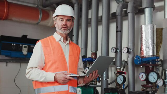 Qualified industrial engineer in safety orange vest and white hard hat using wireless laptop in water treatment hall. Grey bearded man smiling at camera while providing check up at modern workplace.