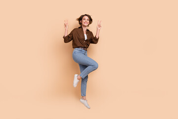 Full length photo of cheerful pretty lady wear brown shirt jumping high showing v-signs empty space isolated beige color background