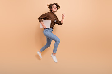Fototapeta na wymiar Full length photo of cute purposeful lady wear brown shirt jumping high holding device running empty space isolated beige color background