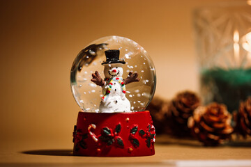 Christmas background 2024. Atmospheric photo of glass snow globe with snow and snowman. New Year brown background. Sphere crystal - a gift for kids. Wintertime holidays fun concept. Warm lights.