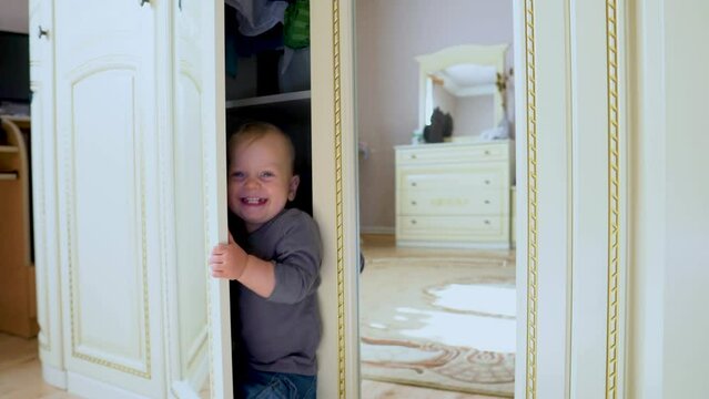 Joyful Caucasian baby with blond hair and blue eyes. Happy funny little boy playing hide and seek at home in a closet room. Child at home. Happy family