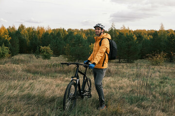 a sporty lifestyle.A man in a protective helmet and equipment stands next to a mountain bike and enjoys nature in a field in autumn