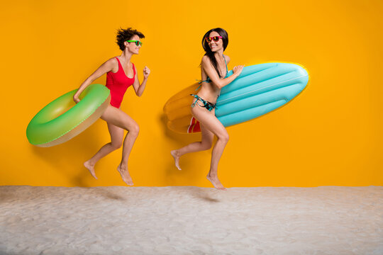 Photo of two charming girls having fun running along coastline swimming sea inflatable toys isolated on vibrant yellow color background