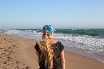 A woman with long blond hair walks along the sandy seashore on a windy day. Travel and tourism.