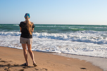 A woman with long blond hair walks along the sandy seashore on a windy day. Travel and tourism.