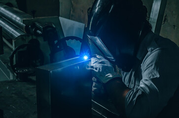 Welder in a protective mask welds a metal box in a locksmith's workshop. Bright light of argon...