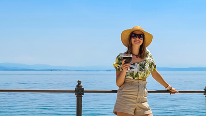 A woman in big sunglasses and a straw hat holding a glass full of dark beer. In the back there is a...