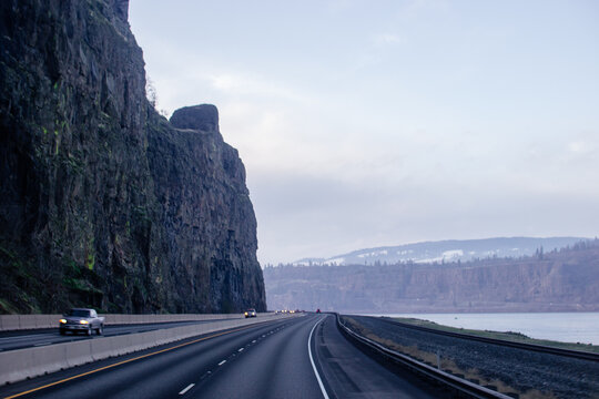 A highway surrounded by high mountains, along which cars drive with their headlights on at dusk